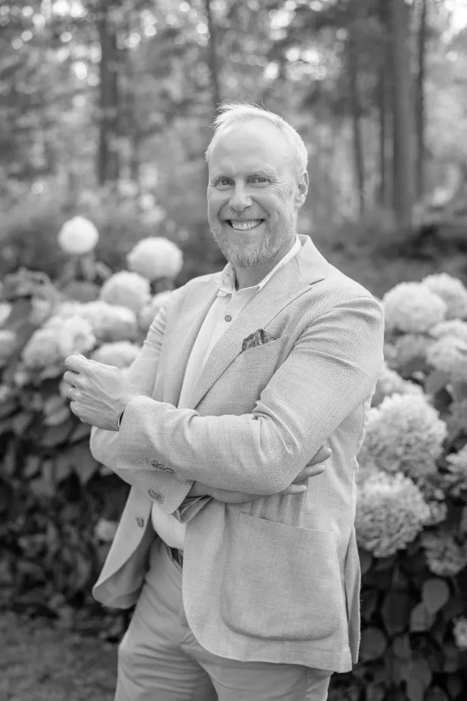 Hymyilevä mies beigeissä vaatteissa seisoo puutarhassa hortensioiden edessä, aurinkoisessa säässä.